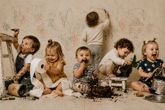 Five toddlers playfully making a mess together in a warm, neutral studio setting, with dirt, paper, and toys scattered around