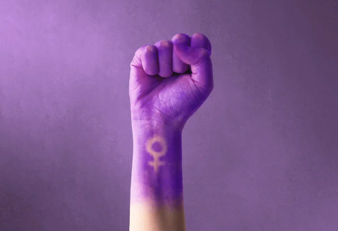 Raised fist painted purple with a Venus symbol on the wrist against a soft purple background, symbolizing feminine strength, power, and empowerment.