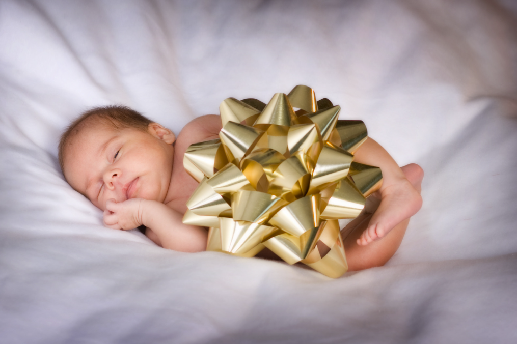 Sleeping Aurelia baby curled beside an oversized gold gift bow on soft white bedding.