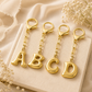 Four gold letter charms spelling ABCD, each attached to a short gold chain and lobster clasp, arranged on a cream fabric surface with dried white flowers around them.
