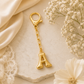 A gold initial keychain charm featuring the letter 'A' in a polished serif font, attached to a short gold chain and lobster clasp. The keychain is styled on a textured stone slab with cream satin fabric, lace, and white baby's breath flowers.