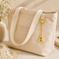 A cream-colored canvas tote bag with a gold initial 'A' charm, styled on a beige surface with a vase of baby's breath, a book with sunglasses, and a soft fabric.