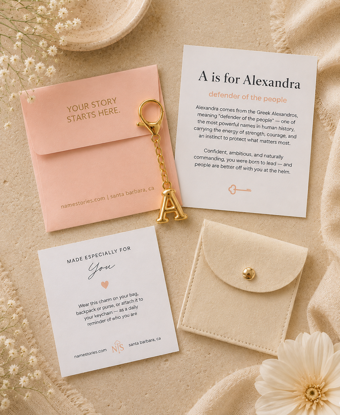 A styled flat-lay of a custom invitation suite from Namestories, featuring a blush pink envelope with gold foil text, a white invitation card with elegant typography, a matching 'Made Especially For' card, and a decorative gold A initial keychain, all arranged on a textured beige fabric background with dried flowers.