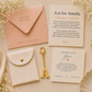 A flat-lay of personalized stationery and accessories on a soft beige fabric background with dried flowers. The set includes a peach envelope with gold text, a cream card reading 'A is for Amelia,' a cream message card, a cream pouch with a gold snap and gold letter A charm, and a gold initial key.