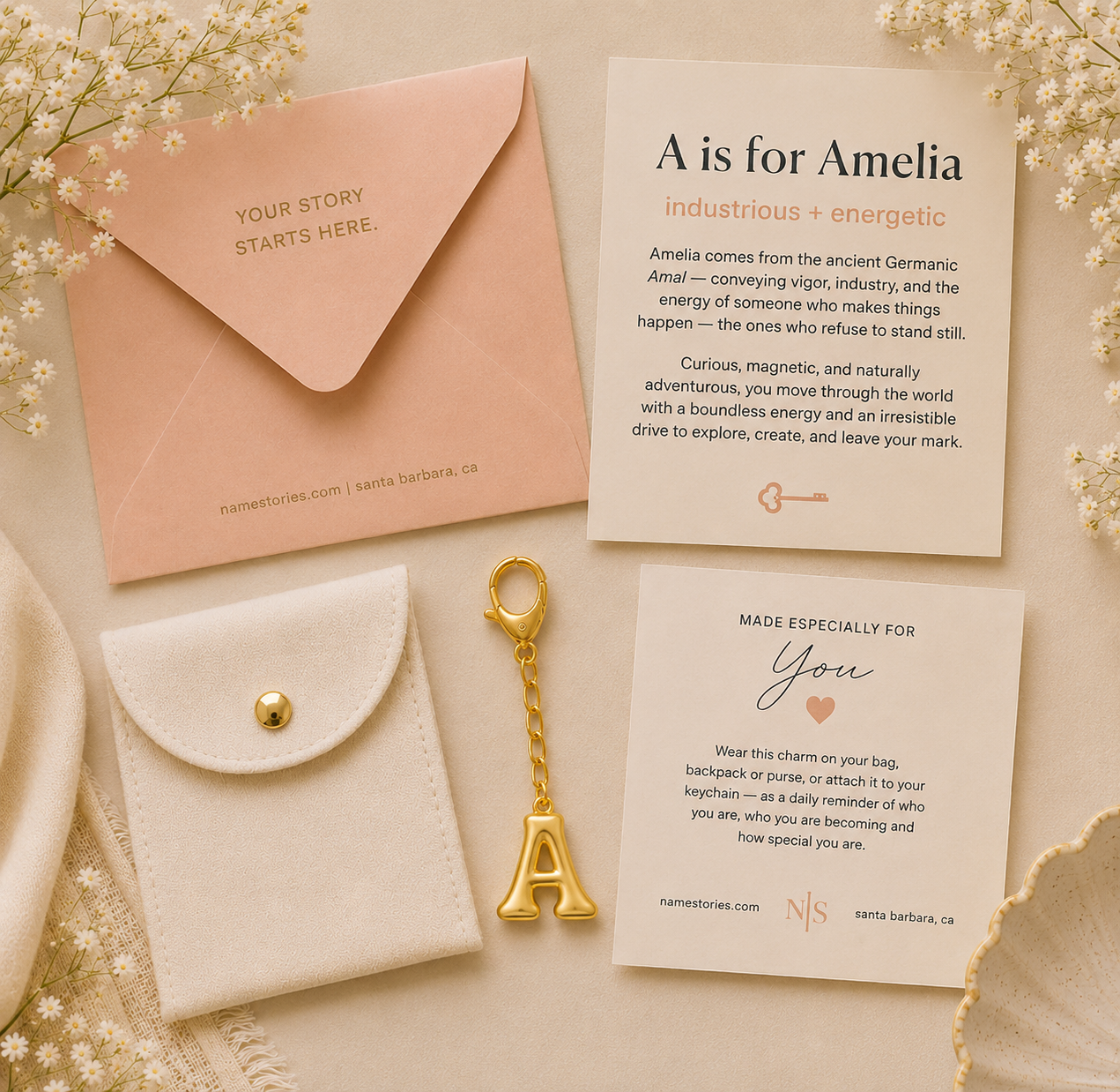 A flat-lay of personalized stationery and accessories on a soft beige fabric background with dried flowers. The set includes a peach envelope with gold text, a cream card reading 'A is for Amelia,' a cream message card, a cream pouch with a gold snap and gold letter A charm, and a gold initial key.
