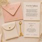 A styled flat-lay of a personalized key gift set on a cream fabric background, featuring a pink envelope, a white card, a white pouch with a gold clasp, a gold key-shaped letter 'A' charm, and a small gold key.