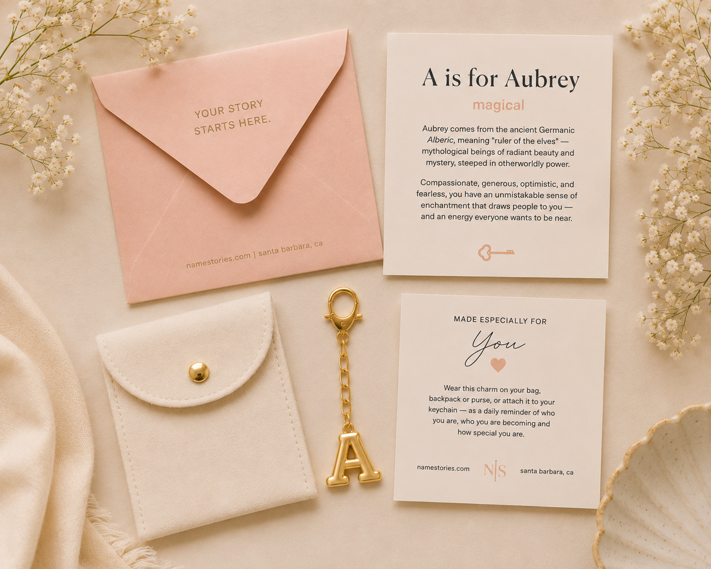 A styled flat-lay of a personalized key gift set on a cream fabric background, featuring a pink envelope, a white card, a white pouch with a gold clasp, a gold key-shaped letter 'A' charm, and a small gold key.