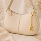 A cream-colored pebbled leather crescent-shaped shoulder bag with gold-tone hardware, featuring a hanging gold 'C' charm, set against a soft beige background with dried flowers.