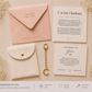 A keepsake gift set arranged on a light beige background with dried floral decorations. The set includes a pale pink envelope with gold text, a white card reading 'C is for Charlotte', a white leatherette pouch with a gold snap, a gold keychain, and another white card.