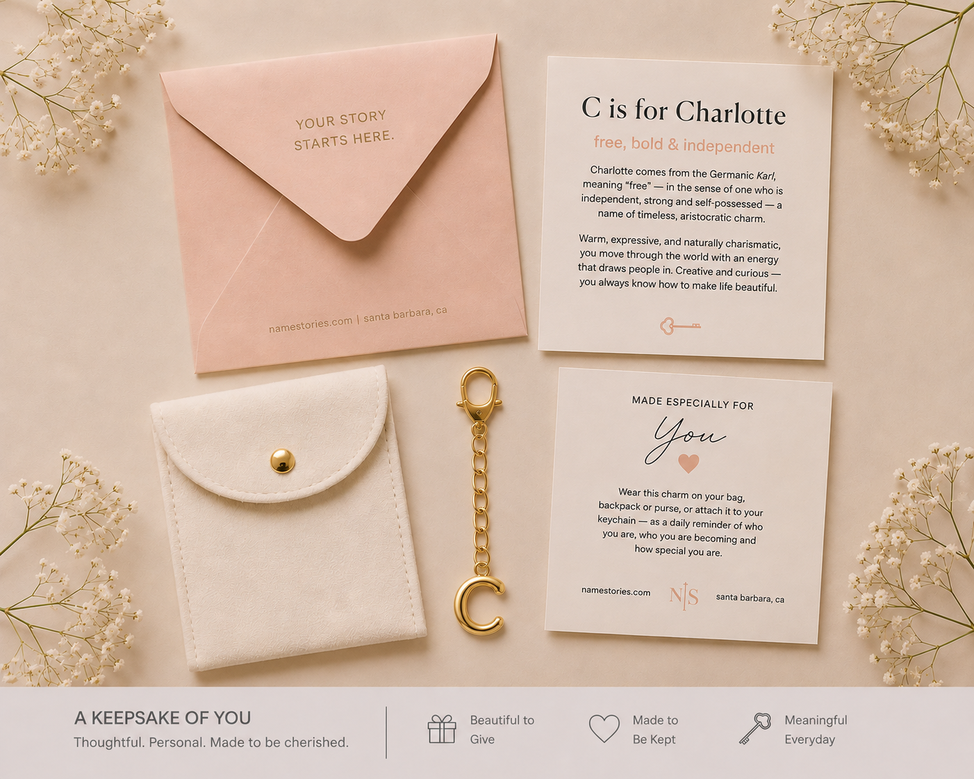 A keepsake gift set arranged on a light beige background with dried floral decorations. The set includes a pale pink envelope with gold text, a white card reading 'C is for Charlotte', a white leatherette pouch with a gold snap, a gold keychain, and another white card.