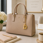 A structured beige handbag with gold-tone hardware and a hanging gold letter D charm, displayed on a desk with a notebook, pen, flowers, and a coffee cup in a bright, styled interior.