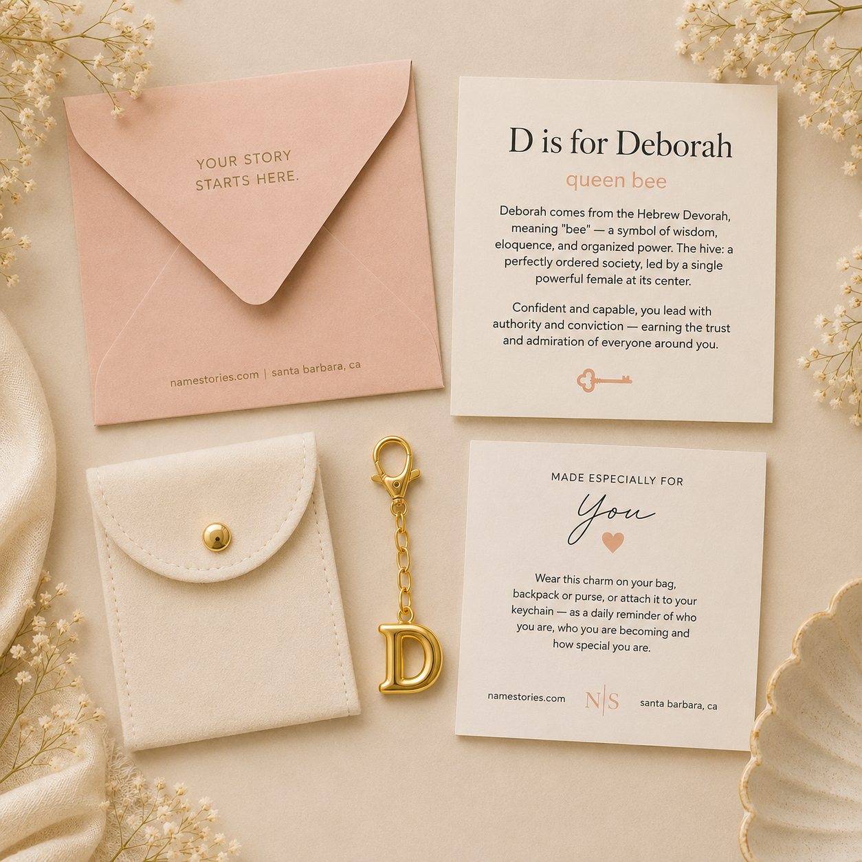A styled flat-lay of a personalized keychain gift set on a cream fabric background with dried flowers. The set includes a blush pink envelope with gold foil text, a white faux-leather card holder with a gold snap, a gold keychain with a decorative letter D charm, and two white printed cards with personalized text.