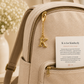 Close-up of a beige canvas backpack with gold-tone hardware, featuring a hanging gold letter K charm and a small white card tucked into the front zip pocket.