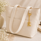 A cream canvas tote bag with dual shoulder straps and a gold hanging initial charm reading 'K', styled on a light beige surface with dried flowers, a vase, a book, and sunglasses nearby.