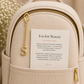 Close-up of a beige backpack with gold-tone hardware, featuring a hanging gold 'S' charm and a white motivational card in the front pocket that reads 'S is for Stacey'.