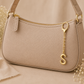 A beige pebbled leather shoulder bag with gold-tone hardware, featuring a gold zipper, a hanging gold keyring charm with an initial 'S', and a matching strap, styled with dried flowers on a neutral beige background.