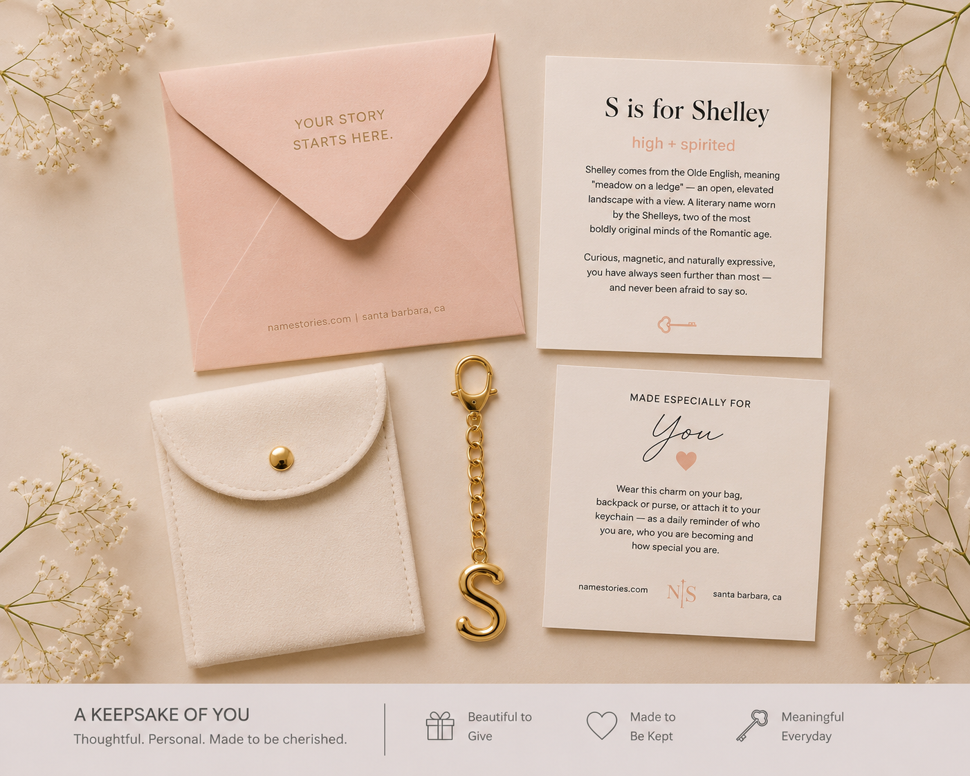 A personalized keepsake gift set laid out on a light beige background with delicate dried flowers. The set includes a pale pink envelope with gold foil text, a cream card with printed text, a white envelope-style pouch with a gold snap and center pin, a gold chain with an initial 'S' keychain, and a printed Shelley keepsake card.