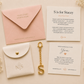 A styled flat-lay of a custom bridal shower gift set on a warm cream background, featuring a pink envelope, a white faux-leather pouch with a gold clasp, a gold keychain with an 'S', and two printed cards.